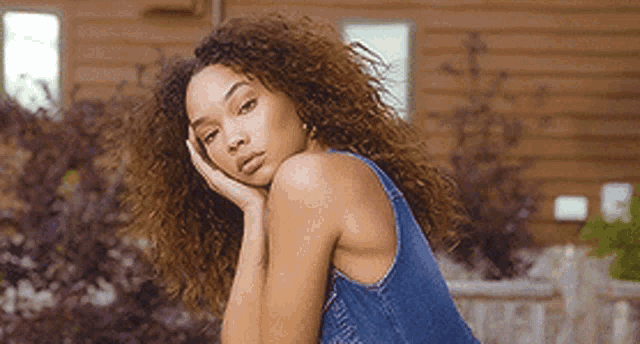 a woman with curly hair wearing a blue denim dress is standing in front of a wooden building .