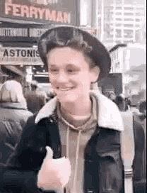 a young man wearing a hat and jacket is giving a thumbs up in front of a ferryman sign .