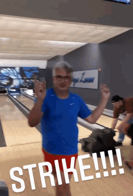 a man in a blue shirt and red shorts stands in a bowling alley with the words strike written on the floor