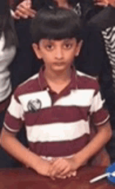 a young boy in a red and white striped shirt is sitting at a table with his hands folded .