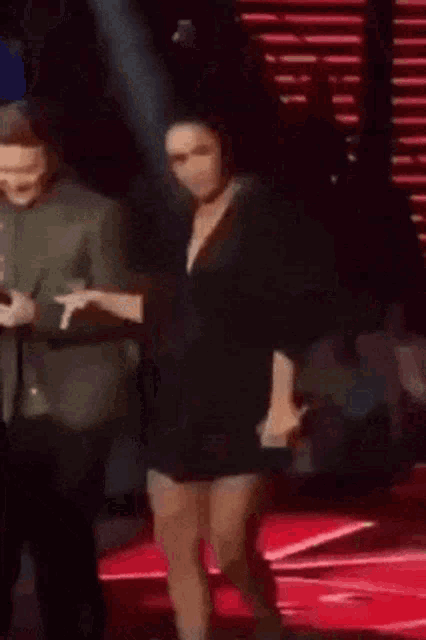 a woman in a black dress is dancing on a stage with a man in a suit behind her