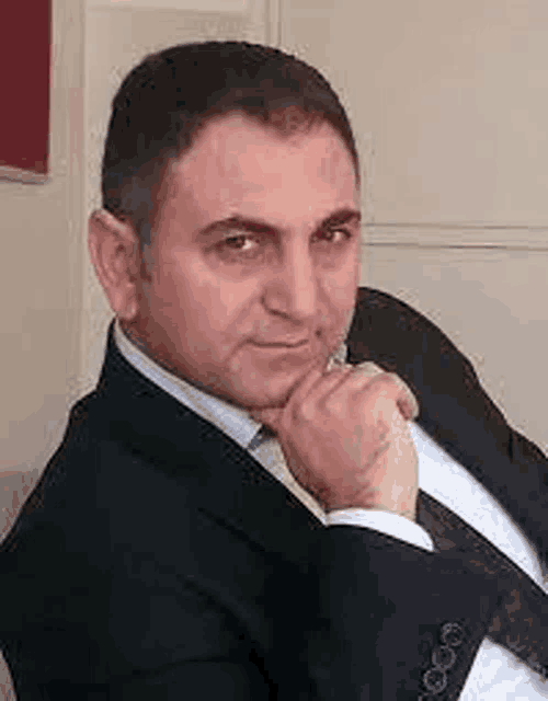 a man in a suit and tie is sitting down with his hand on his chin and looking at the camera .