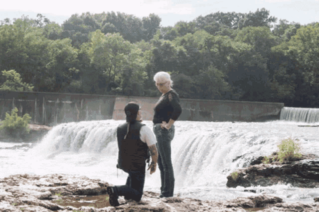 a man is kneeling down in front of a waterfall to propose to a woman