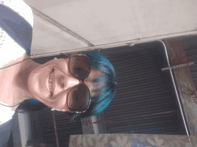 a woman with blue hair and sunglasses is smiling in front of a sign that says downstairs