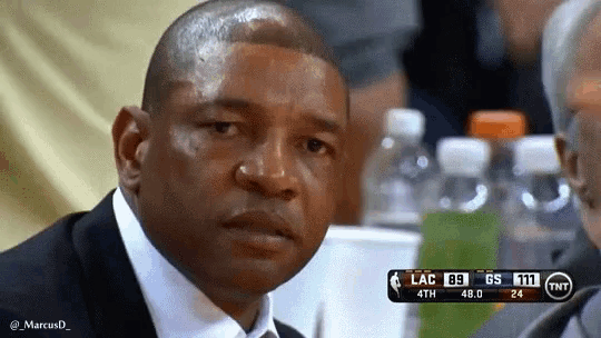 a man in a suit and tie is sitting at a table watching a basketball game with a scoreboard behind him .