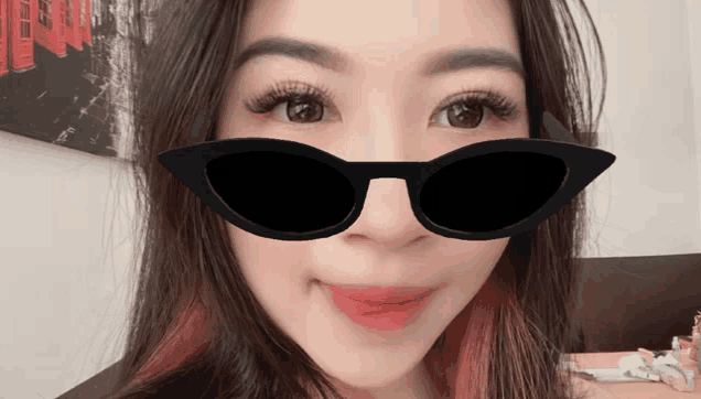 a woman wearing sunglasses looks at the camera in front of a painting that says ' a ' on it