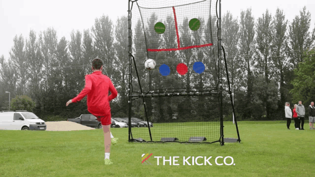 a boy in a red jacket is kicking a soccer ball in front of a goal that says the kick co. on it