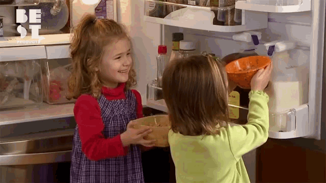 two little girls are standing in front of an open refrigerator . one of the girls is holding a bowl of food .