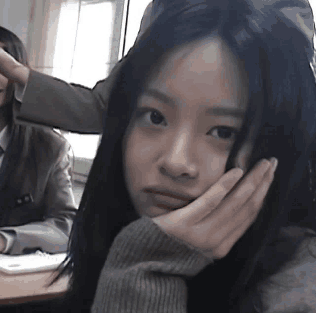 a girl with long black hair is sitting at a desk with her hand on her chin