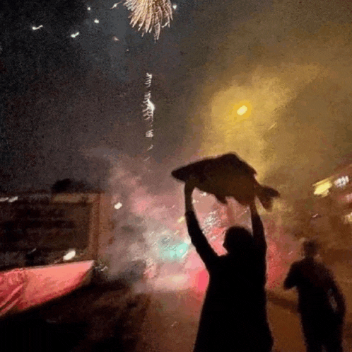 a man is holding a large fish in the air in front of fireworks