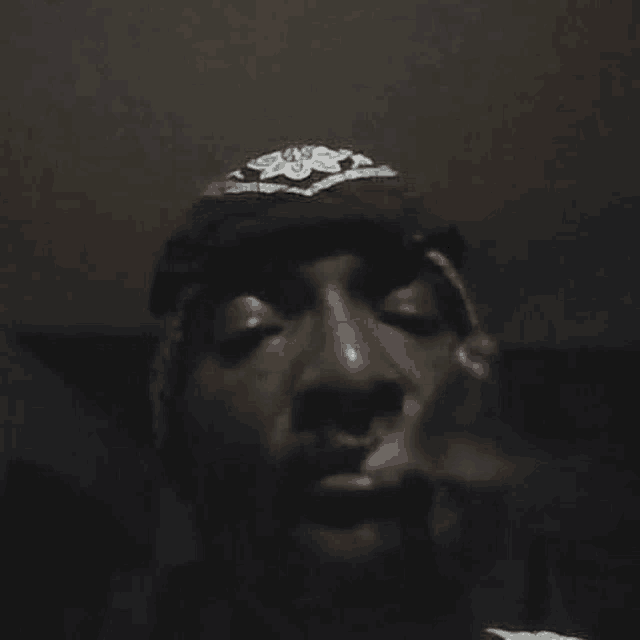 a man wearing a hat and a bandana is smoking a cigarette in a dark room .