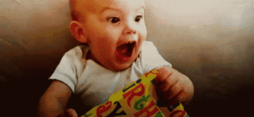 a baby is sitting on a couch with his mouth open and holding a piece of birthday wrapping paper .