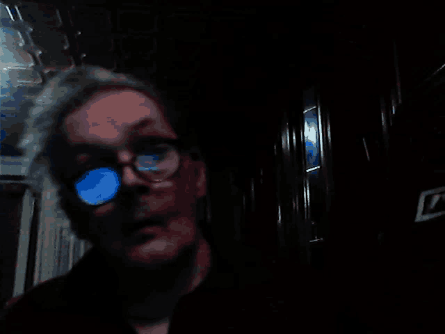 a man wearing glasses looks at the camera in a dark hallway