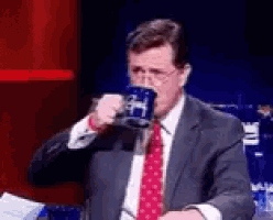 a man in a suit and tie is drinking from a mug with the letter h on it .