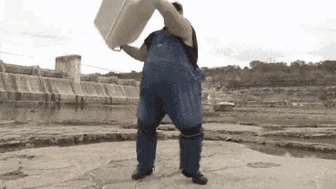 a man in blue overalls is holding a white bucket over his head while standing in front of a body of water .