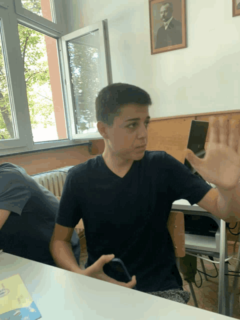 a boy in a black shirt is sitting at a desk with his hand out