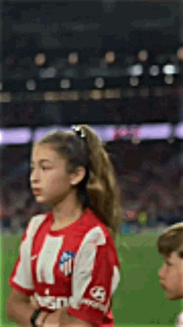 a young girl wearing a red and white striped shirt is standing on a soccer field with her eyes closed .