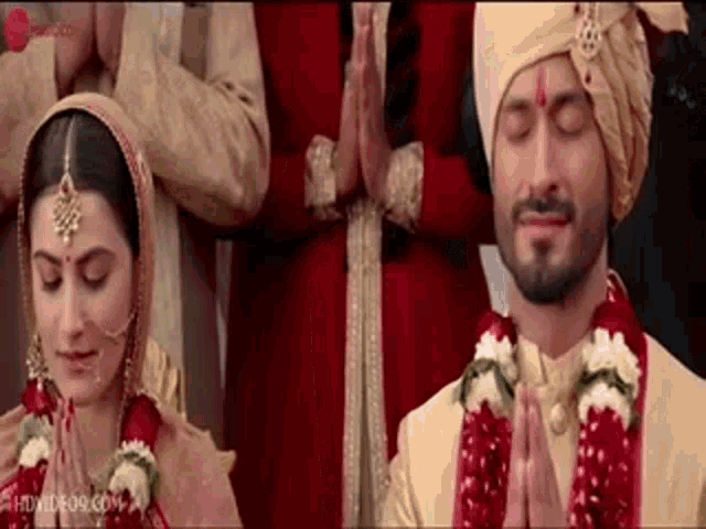 a bride and groom are standing next to each other with their eyes closed and their hands folded in prayer .