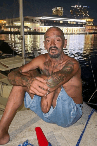 a man with a tattoo on his chest sits on a dock in front of a body of water