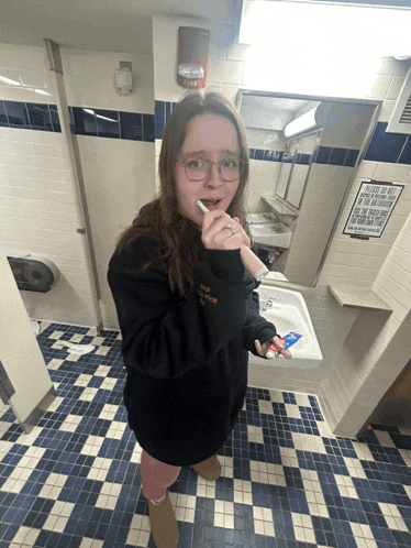 a girl brushing her teeth in a bathroom with a sign on the wall that says wash your hands before using the toilet