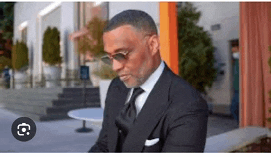 a man in a suit and tie is sitting in front of a building wearing sunglasses .