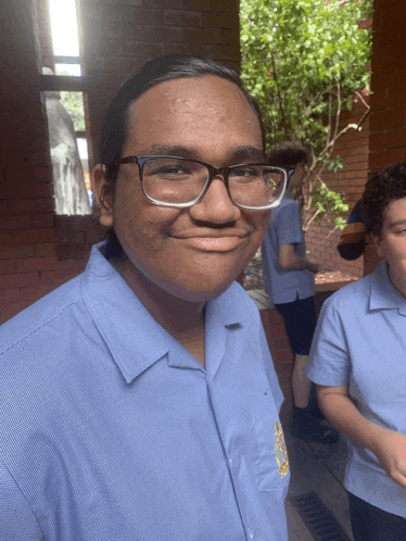 a young man wearing glasses and a blue shirt with a yellow emblem on the sleeve smiles for the camera