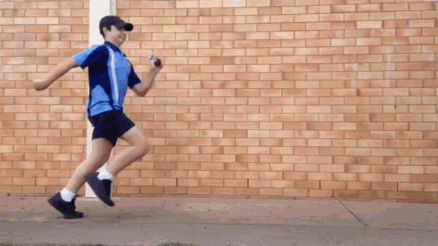 a boy in a blue shirt and black shorts is running on a sidewalk in front of a brick wall