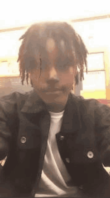 a young man with dreadlocks is taking a selfie while wearing a black jacket and white shirt .