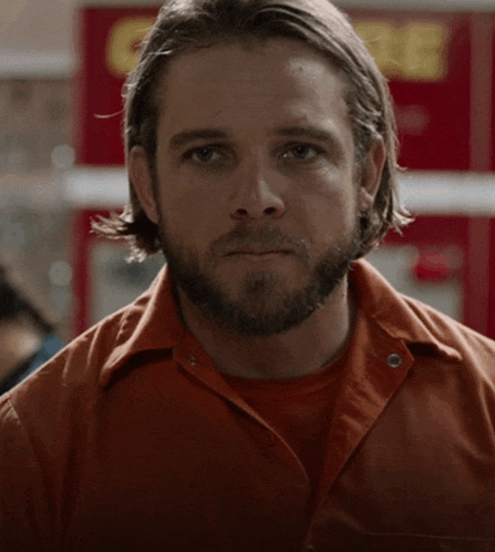 a man with a beard wearing an orange shirt is standing in front of a store that says coca cola