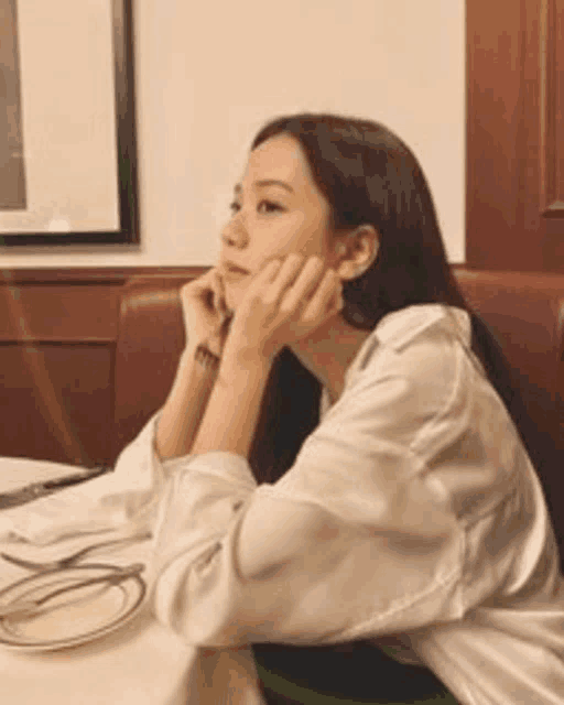 a woman in a white shirt is sitting at a table with her hands on her chin .