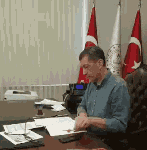 a man sits at a desk in front of three flags including one that says ' turkey ' on it