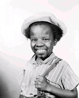 a black and white photo of a young boy wearing a hat and suspenders and smiling .