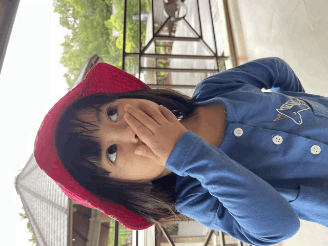 a little girl wearing a blue shirt and a red hat is covering her mouth with her hand