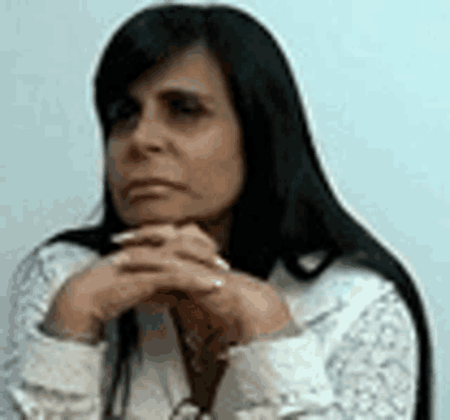 a woman with long black hair is sitting in front of a white wall with her hands folded on her chin .