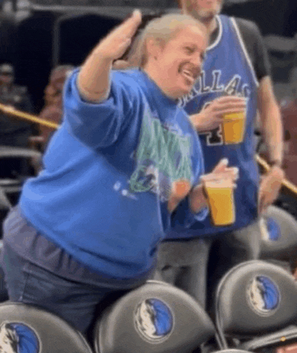 a woman in a dallas jersey is sitting in the stands at a basketball game holding a cup of beer .