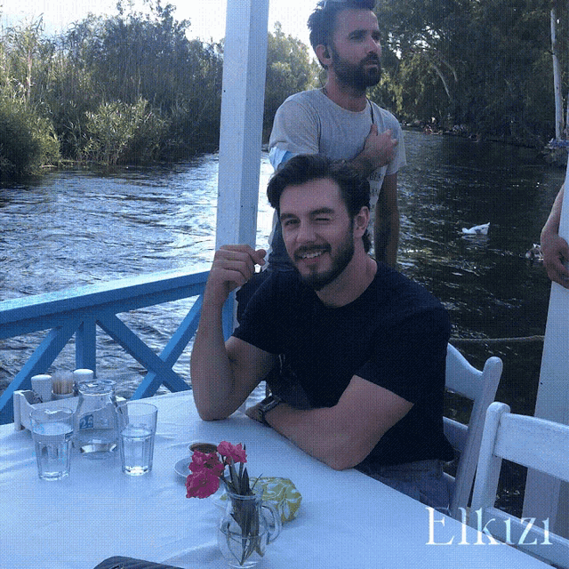 a man with a beard sits at a table in front of a river with the word elkizi on the bottom