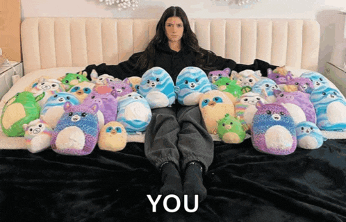 a woman is sitting on a bed surrounded by stuffed animals with the words " you " above her