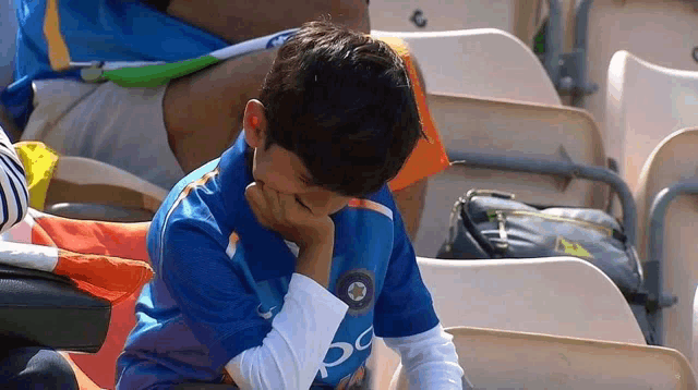 a young boy in a blue shirt is sitting in a stadium with his hand on his face .