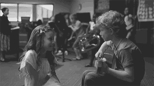 a boy and a girl are looking at each other in a black and white photo . the boy is playing a guitar .