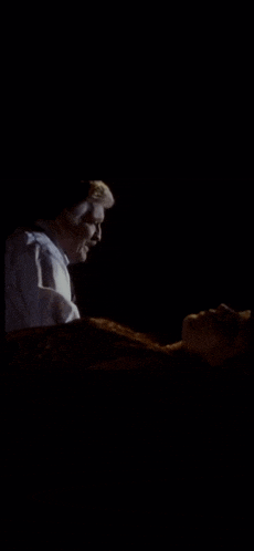 a man is standing next to a woman who is laying on a bed in the dark .