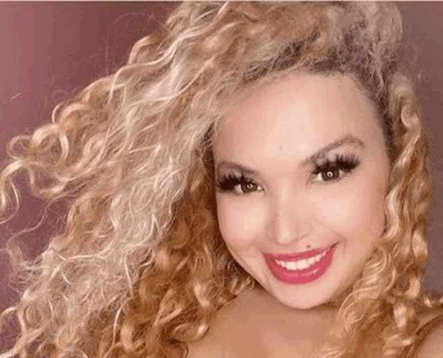 a woman with blonde curly hair is smiling and wearing pink lipstick .