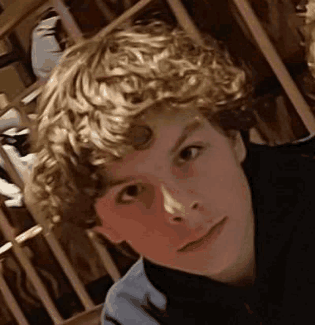 a young boy with curly hair is standing in front of a wooden shelf and looking at the camera .