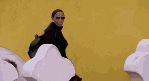 a woman wearing sunglasses and a black turtleneck sweater is walking in front of a yellow wall .
