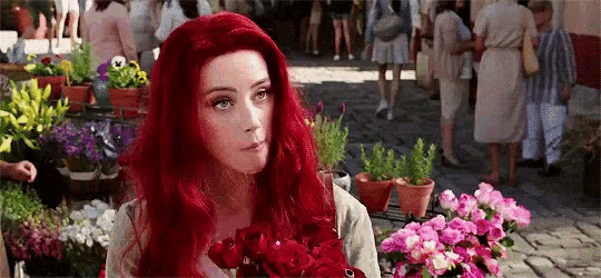 a woman with red hair is holding a bouquet of red roses in front of a flower stand .