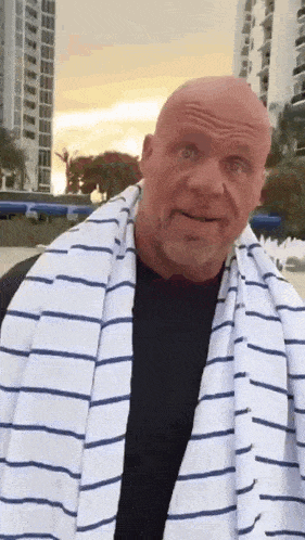 a bald man wearing a striped towel around his neck is standing in front of a building .