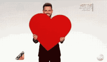 a man in a suit and tie is sitting on a white couch holding a microphone and a sign that says meet the bachelor