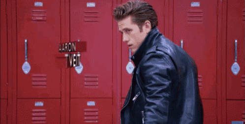 a man in a leather jacket is standing in front of red lockers with the name aaron tveit written above him