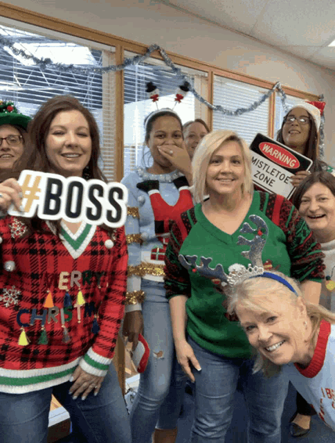 a group of women wearing ugly christmas sweaters are posing for a picture and one woman is holding a sign that says boss