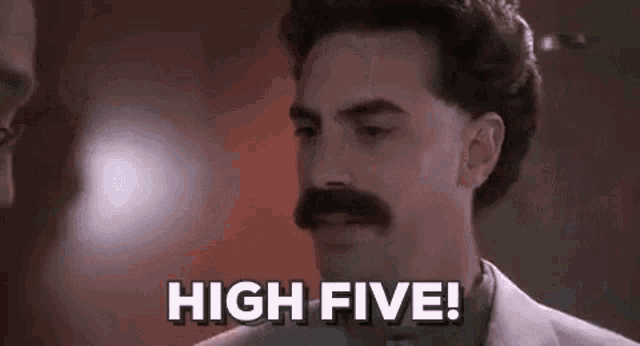 a man with a mustache is standing in front of a wall and says `` high five '' .