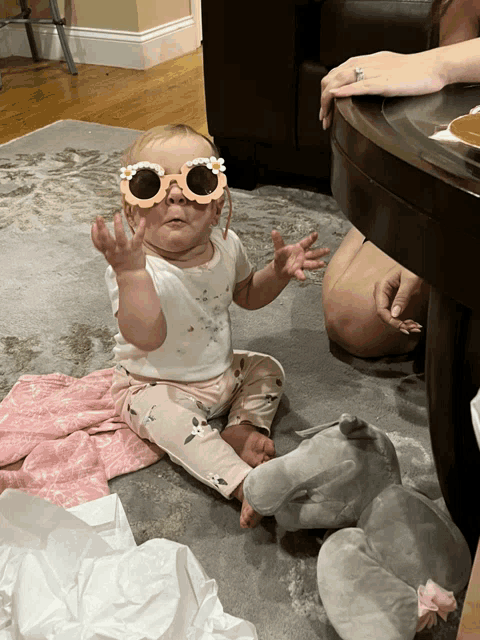 a baby wearing sunglasses sits on the floor next to a stuffed animal